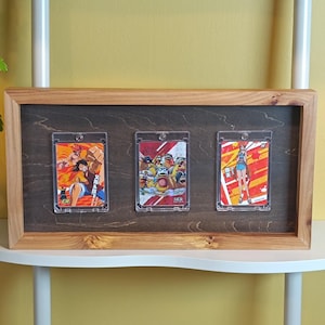 Peut inclure: Un cadre de présentation rectangulaire en bois avec un panneau arrière teinté foncé contenant trois étuis à cartes en plastique transparent. Chaque étui contient une carte à collectionner anime colorée avec des personnages illustrés sur des fonds graphiques orange, rouge et jaune.