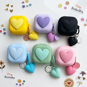 Può includere: Sei custodie quadrate in silicone per auricolari nei colori giallo, lavanda, nero, azzurro, verde salvia e rosa. Ogni custodia presenta un cuore in rilievo sul davanti e un ciondolo a forma di cuore abbinato con un piccolo smiley circolare.