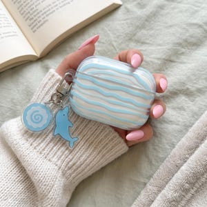 Puede incluir: Una funda protectora transparente para auriculares inalámbricos con rayas horizontales onduladas de color azul claro. La funda incluye un clip de metal plateado con dos dijes acrílicos: uno circular con un patrón de espiral y un delfín azul.
