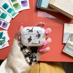May include: A clear protective case for wireless earbuds featuring a black and white cat and fish pattern. The case includes a decorative beaded charm strap with black and white beads, a black star-shaped bead, and a black cat-shaped charm.