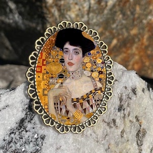 May include: A gold-toned oval brooch with a detailed ornate frame. The brooch features a portrait of a woman with dark hair and a pearl necklace, set against a background of gold and black geometric patterns.