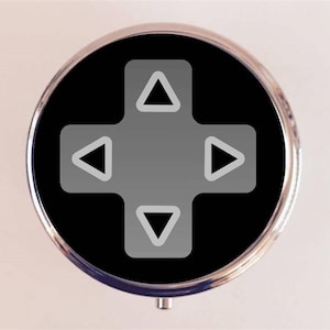 May include: A round, silver-colored pill box with a black background and a white directional pad icon. The icon has four arrows pointing up, down, left, and right.