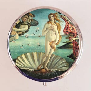 May include: A round, silver-toned compact mirror featuring a print of Sandro Botticelli's "The Birth of Venus." The artwork depicts Venus standing in a seashell, with figures on either side. The background shows a seascape with a blue sky.