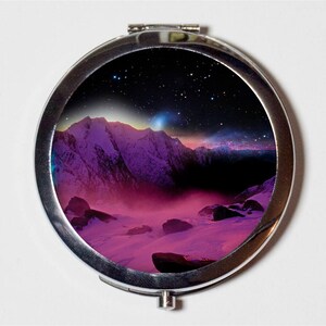 May include: A silver compact mirror with a colorful image of a mountain range under a starry night sky. The mountains are covered in snow and the sky is a gradient of pink, purple, and blue.