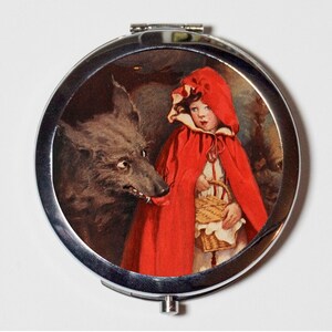 May include: A silver compact mirror with a vintage illustration of Little Red Riding Hood and a wolf. The girl is wearing a red hooded cloak and holding a basket.
