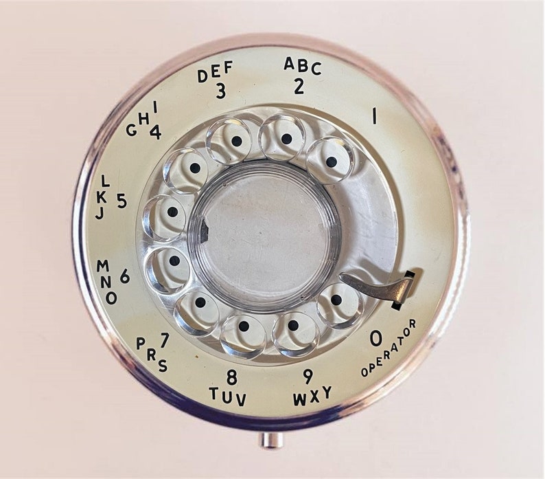 Puede incluir: Un tel&eacute;fono de disco rotatorio vintage con una cara blanca y letras negras. El dial tiene los n&uacute;meros del 2 al 9 y las letras ABC al WXY. El dial tambi&eacute;n tiene un 0 para el operador.