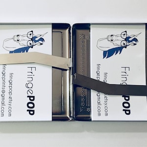 Puede incluir: Una cajetilla de cigarrillos de metal plateado con una tarjeta blanca en su interior. La tarjeta presenta un unicornio de dibujos animados con gafas y el texto "Fringe POP" con informaci&oacute;n de contacto.
