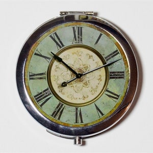 May include: A silver compact mirror with a vintage clock face design. The clock face is green and white with Roman numerals and black hands. The clock face has a distressed look.