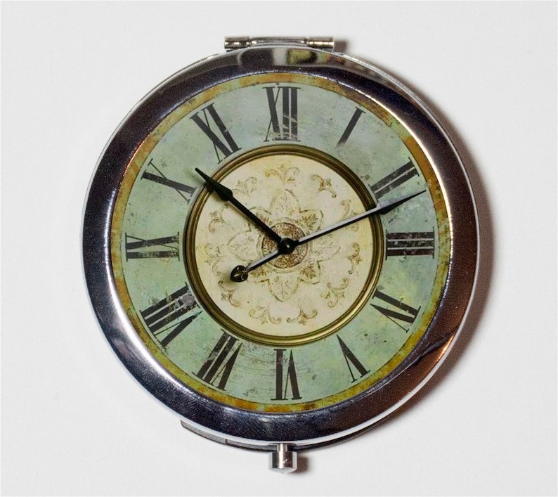 May include: A silver compact mirror with a vintage clock face design. The clock face is green and white with Roman numerals and black hands. The clock face has a distressed look.