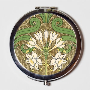 May include: A silver compact mirror with a floral design. The design features white flowers with yellow centres and green leaves on a brown background.