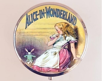 May include: A round, silver-colored compact with a colorful image of Alice from Alice in Wonderland. The image shows Alice in a white dress with a pink skirt and a white apron. She is looking down at a white rabbit with a blue vest and a pocket watch. The text "Alice-in-Wonderland" is printed in blue and white letters around the image.