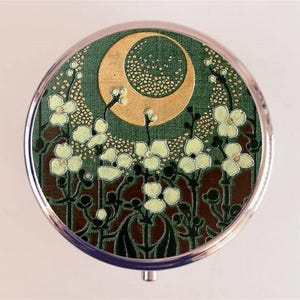 Könnte beinhalten: Runder silberner Kompaktspiegel mit grünem und goldenem Blumendesign. In der Mitte befindet sich ein goldener Halbmond und weiße Blumen vor einem grünen Hintergrund. Das florale Design ist vor einem dunkelbraunen Hintergrund.