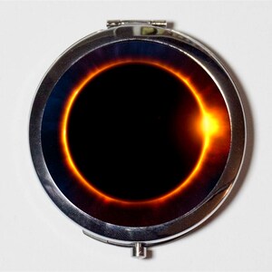 May include: A silver compact mirror with a black and orange image of a solar eclipse.