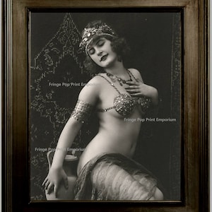 May include: A black and white vintage photograph of a woman wearing a jeweled headdress and a beaded bra. She is sitting on a chair with a patterned fabric background. The image is framed in a dark wood frame.