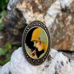 May include: A vintage-style oval brooch with a detailed metal frame. The brooch features a colorful image of a woman in a yellow hat applying lipstick.