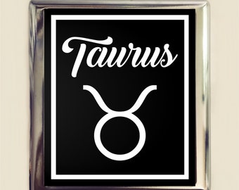May include: A silver metal sign with a black background and white text. The sign says "Taurus" and features the Taurus zodiac symbol.