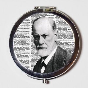 May include: A circular, silver compact mirror with a black and white portrait of Sigmund Freud. The background is a collage of dictionary pages. The compact is open, displaying the image.