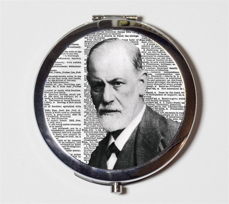 May include: A circular, silver compact mirror with a black and white portrait of Sigmund Freud. The background is a collage of dictionary pages. The compact is open, displaying the image.