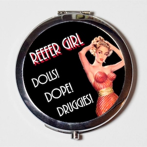 May include: A silver compact mirror with a black background and a red and white image of a woman with the text "REEFER GIRL" in red and "DOLLS! DOPE! DRUGGIES!" in white.