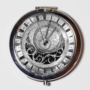 May include: A silver compact mirror featuring a black and white image of an elaborate clock face. The clock face has Roman numerals and a detailed design.