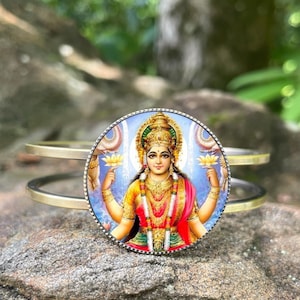 May include: A gold-toned metal bracelet featuring a circular pendant with a colorful illustration of a deity. The image shows a woman in a red and yellow sari holding lotus flowers. The bracelet rests on a textured gray rock.
