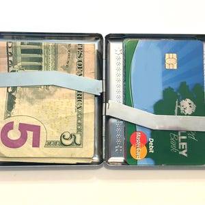 Puede incluir: Una cajetilla de cigarrillos de metal plateado con un billete de 5 d&oacute;lares y una tarjeta de cr&eacute;dito en su interior. La tarjeta de cr&eacute;dito es una tarjeta de d&eacute;bito de Valley Bank y tiene el logotipo de Mastercard. La cajetilla tiene un cierre de metal.