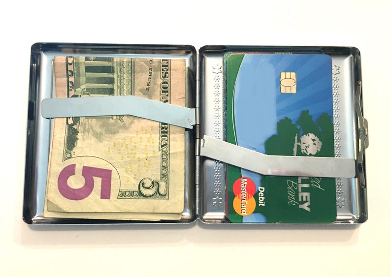 Puede incluir: Una cajetilla de cigarrillos de metal plateado con un billete de 5 d&oacute;lares y una tarjeta de cr&eacute;dito en su interior. La tarjeta de cr&eacute;dito es una tarjeta de d&eacute;bito de Valley Bank y tiene el logotipo de Mastercard. La cajetilla tiene un cierre de metal.