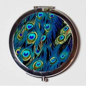 May include: A silver compact mirror with a peacock feather design. The design features blue, green, and gold peacock feathers with eye patterns.