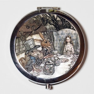 May include: A round, silver compact mirror featuring a detailed illustration of the Mad Hatter's tea party from Alice in Wonderland. The image shows the Mad Hatter, the March Hare, and Alice at a table set with tea cups and a teapot.