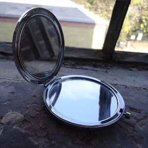 May include: A silver compact mirror with a round, hinged lid. The mirror is open, revealing a reflective surface.