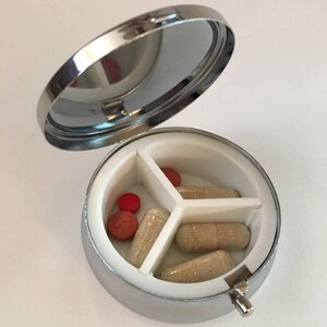 May include: A silver, round pill organizer with a hinged lid, open to reveal three compartments. The compartments contain various pills and capsules in shades of red and beige. The interior is white.