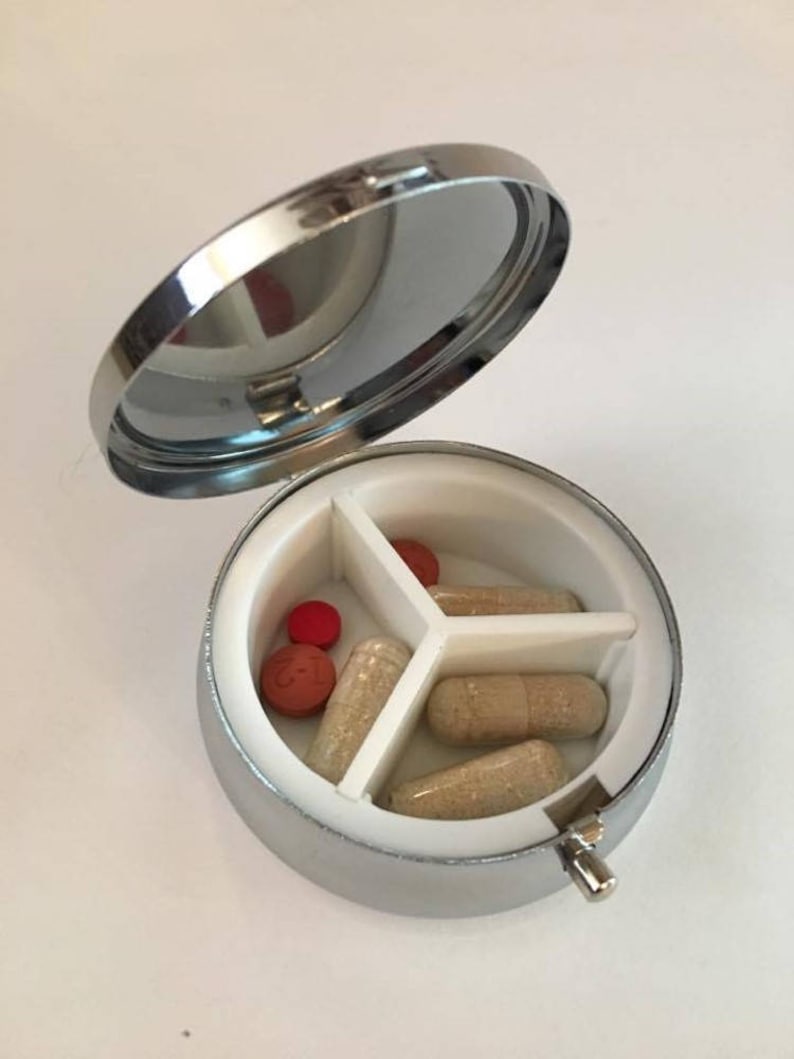 May include: A silver, round pill organizer with a hinged lid, open to reveal three compartments. The compartments contain various pills and capsules in shades of red and beige. The interior is white.