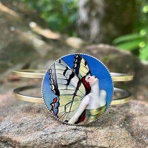 May include: A gold-toned metal cuff bracelet featuring a round, blue-backed image of a woman with butterfly wings. The woman has red hair and is looking upwards. The butterfly wings are white, black, and yellow. The bracelet is resting on a rock.