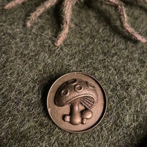 Könnte beinhalten: Eine bronzene Metallstifte mit einem detaillierten Pilzdesign. Der Pilz hat eine gewölbte Kappe mit einer strukturierten Oberfläche und einen Stiel mit einer abgerundeten Basis.