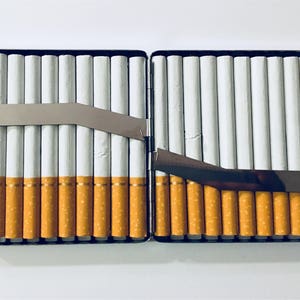 Puede incluir: Una cajetilla de cigarrillos de plata con una tapa con bisagras, abierta para revelar filas de cigarrillos. La cajetilla tiene un clip de metal en el lateral.