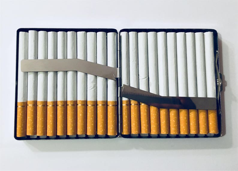 Puede incluir: Una cajetilla de cigarrillos de plata con una tapa con bisagras, abierta para revelar filas de cigarrillos. La cajetilla tiene un clip de metal en el lateral.