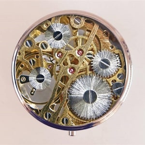 May include: A round, silver-toned, decorative box with a clear top revealing a complex, intricate, gold-toned, clockwork mechanism.
