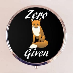 May include: A round, silver-colored compact with a black background. The compact features a cartoon fox with an orange and white fur pattern. The text "Zero Given" is printed in white on the compact.