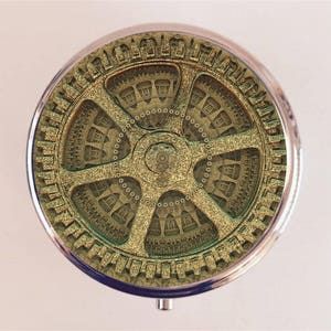 May include: A round, silver-colored trinket box with a gold-colored, intricate, gear-like design on the lid.
