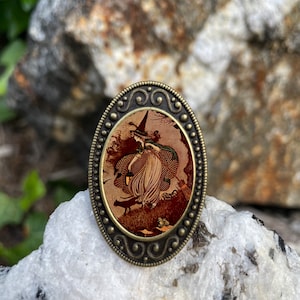 May include: An oval-shaped, bronze-colored pendant with a detailed, ornate border. The pendant features a vintage illustration of a witch flying on a broomstick with a black cat and a lantern.