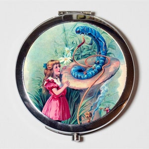 May include: A silver compact mirror with a colorful image of Alice from Alice in Wonderland talking to a blue caterpillar on a mushroom.