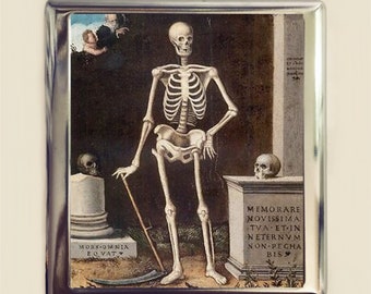 May include: A vintage cigarette case featuring a painting of a skeleton standing in a dark room. The skeleton is holding a scythe and a staff. The painting is surrounded by a silver frame.