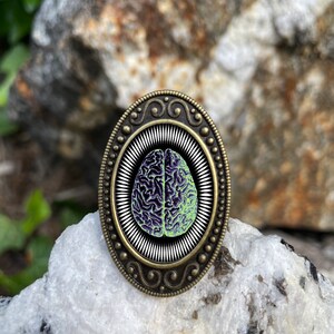 May include: A bronze oval-shaped ring with a detailed design. The ring features a black and white image of a human brain with green accents.