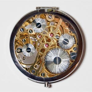 May include: A silver compact mirror with a clear lid revealing a gold-toned intricate clockwork mechanism.