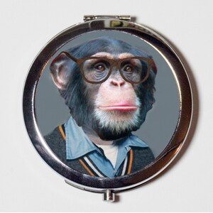 May include: A silver compact mirror with a round, grey background featuring a chimpanzee wearing brown-rimmed glasses and a blue and black sweater.