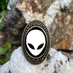 Könnte beinhalten: Eine weiße Alien-Gesichts-Silhouette auf schwarzem Hintergrund, gerahmt in einer ovalen, verzierten, bronzefarbenen Metalleinfassung.