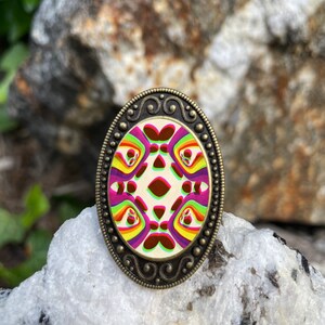 May include: An oval-shaped, bronze-colored ring with a colorful, abstract design. The design features a repeating pattern of pink, yellow, green, and red shapes.