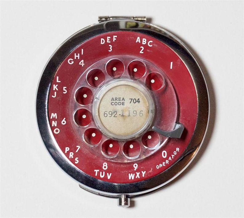 May include: A red rotary phone dial with a silver trim and a clear plastic cover. The dial has the letters ABC, DEF, GHI, JKL, MNO, PRS, TUV, WXY, and 0, as well as the numbers 2, 3, 4, 5, 6, 7, 8, 9, and 0. The dial also has the text "AREA CODE 704 692-1196" and "OPERATOR".