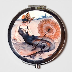 May include: A vintage-style compact mirror with a colorful illustration of a mermaid with an orange and black striped hat and a parasol. The mermaid is lying on a beach with a blue tail.