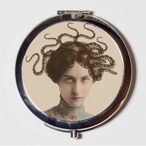May include: A silver compact mirror with a vintage image of a woman with an octopus on her head. The woman has dark hair and is wearing a light-colored blouse.